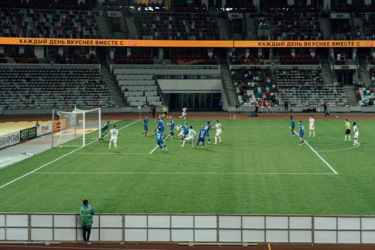 Vista aérea de um estádio de futebol moderno durante uma partida internacional. Jogadores em uniformes amarelos e verdes são vistos em campo, parecendo silhuetas distantes, com uma atmosfera um pouco