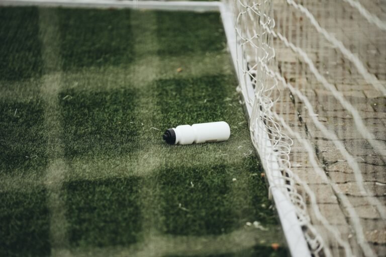 Jogadores de futebol tomando água durante uma pausa em campo sob sol intenso, com torcedores observando no estádio