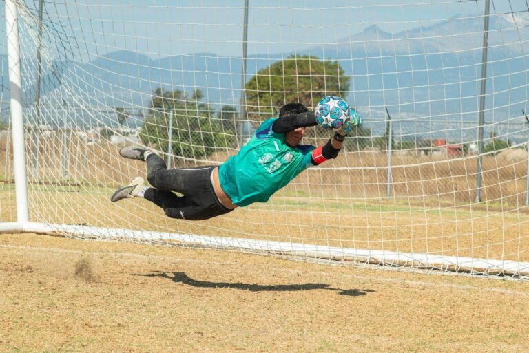 Imagem de um jogador de futebol chutando um pênalti enquanto o goleiro se estende para salvar, com a torcida vibrando no estádio.
