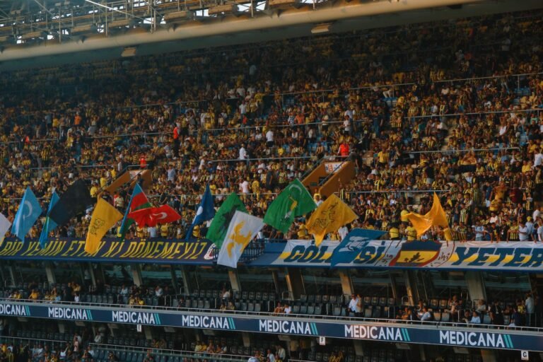Câmera aérea de um estádio de futebol lotado, com torcedores segurando bandeiras e faixas, demonstrando entusiasmo e expressão política durante uma partida.