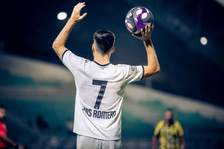 Jogador de futebol celebrando gol em campo com confete e torcida aplaudindo