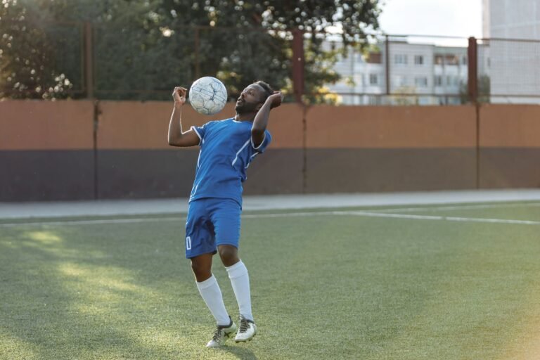 Jovem atacante de futebol treinando com a bola em campo, com estádio ao fundo.