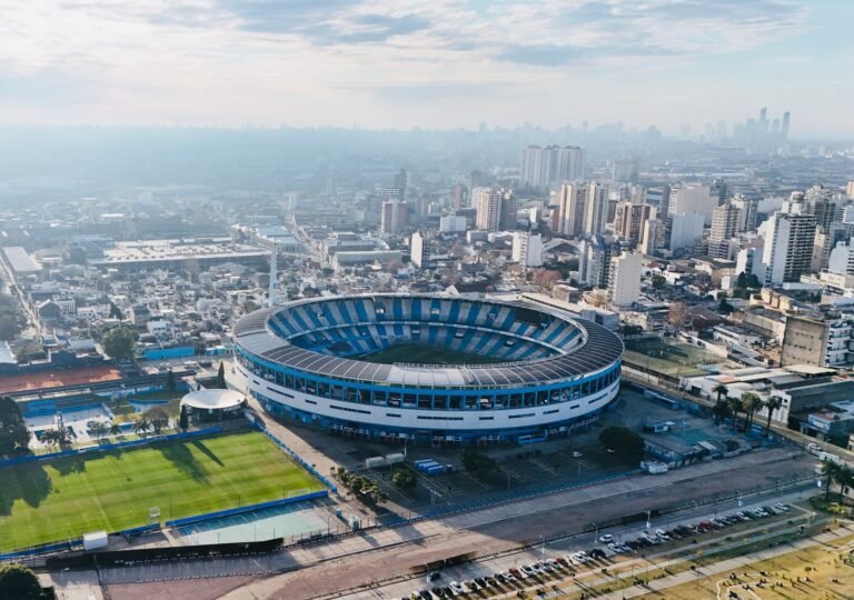Estádio de futebol com bandeiras brasileira e argentina e silhueta de jogador no centro do gramado.