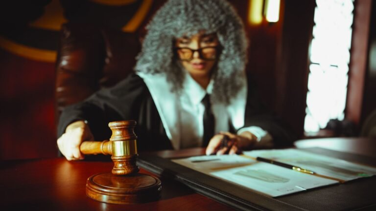 Sala de tribunal com mesa de documentos legais, um troféu de futebol sobre a mesa e silhuetas genéricas de pessoas no fundo.