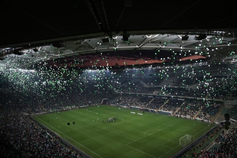 Estádio lotado com palco iluminado e artistas em show de intervalo, torcedores vibrando.