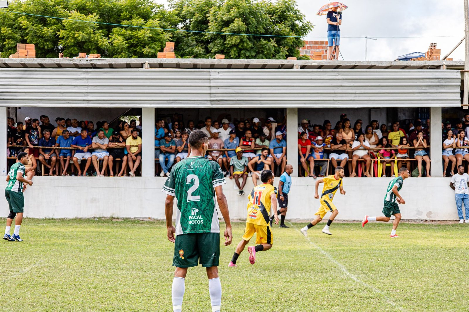 Estádio de futebol à noite com chuva, treinador no banco, jogadores em ação e placar mostrando placar apertado.